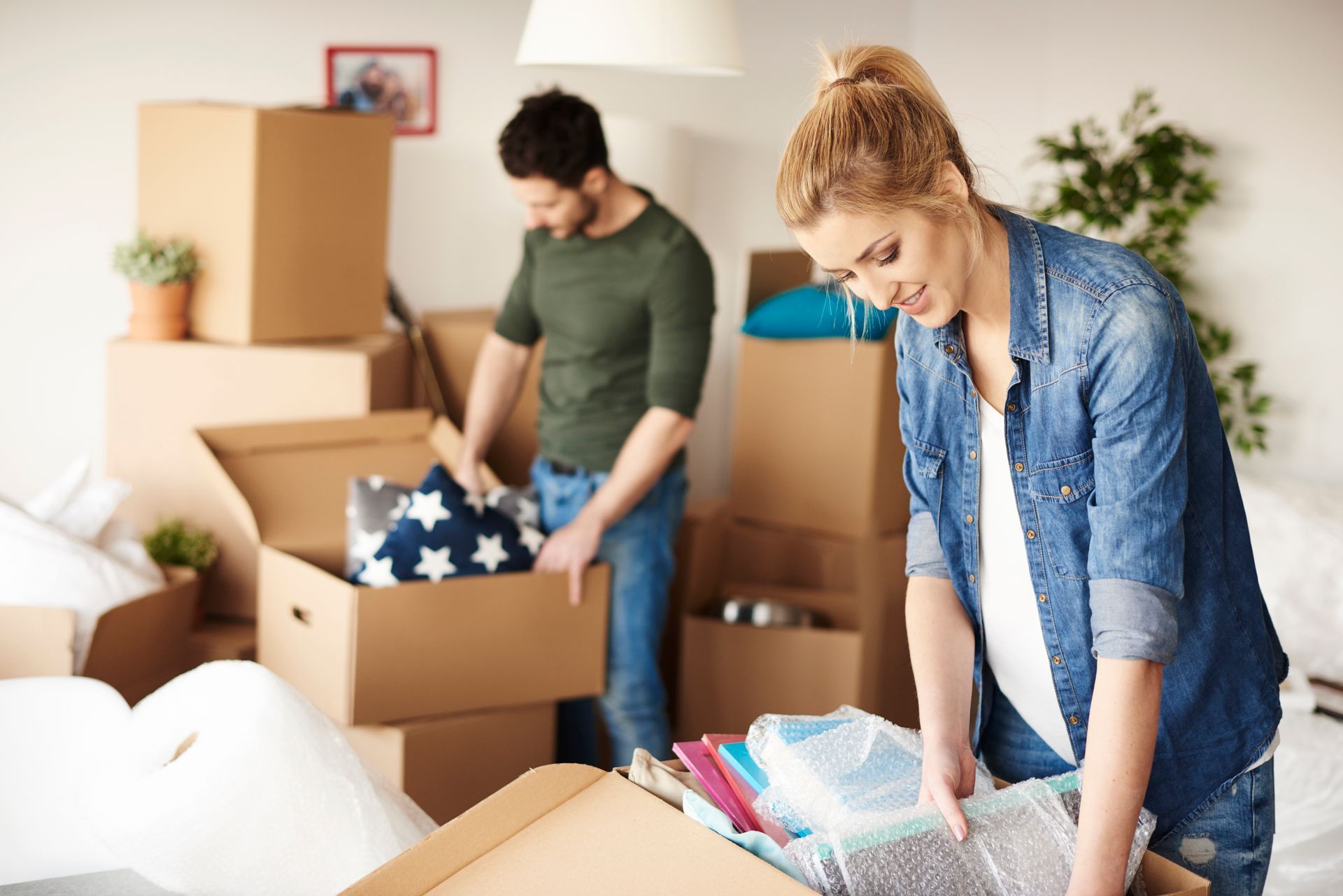 Couple packing boxes in a room filled with moving supplies. Man in green shirt, woman in blue.