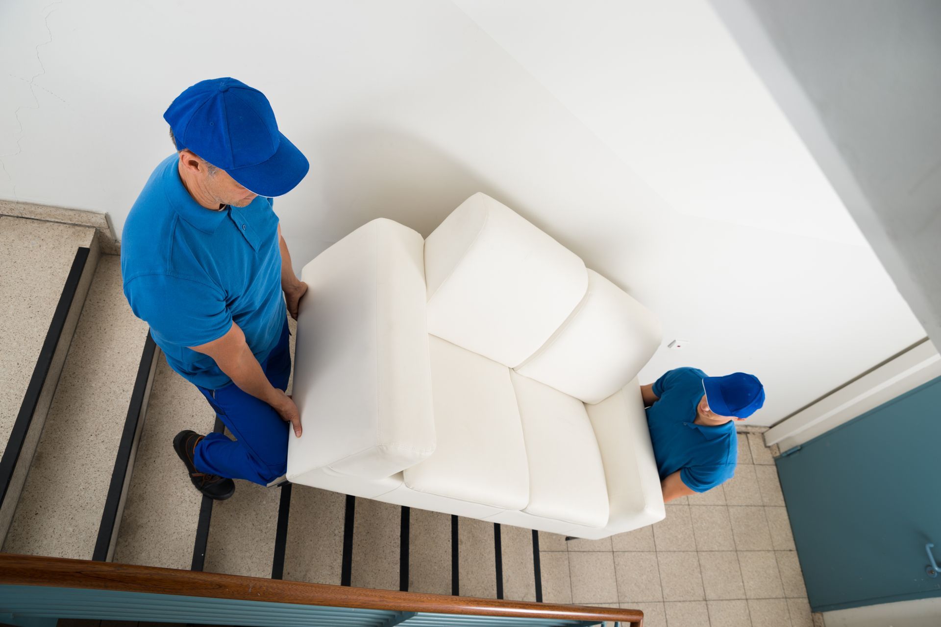 Two movers in blue uniforms carrying a white sofa down stairs.
