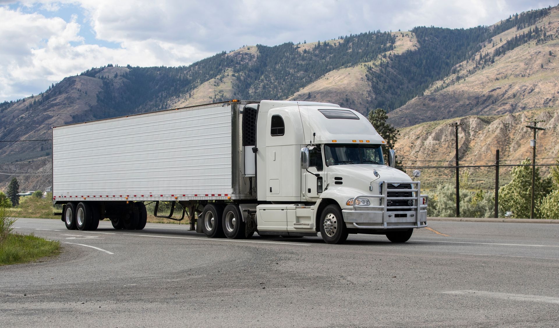 A white semi truck is driving down a road with mountains in the background.