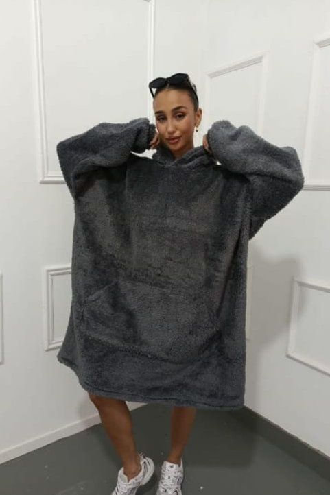 Mujer con una manta con capucha gris de gran tamaño y zapatos blancos en una habitación con ribete cuadrado blanco.
