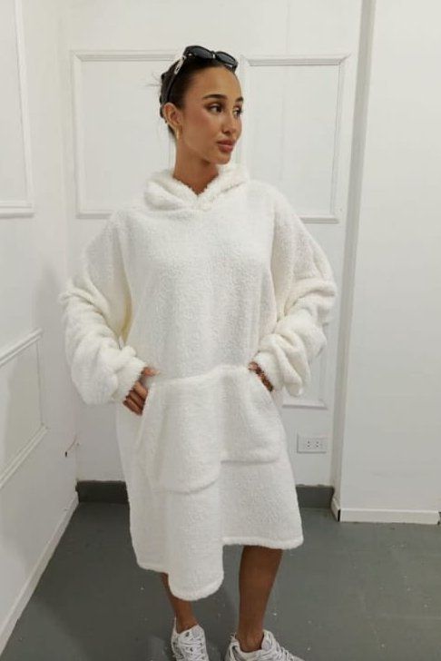 Mujer con vestido blanco esponjoso con capucha, con las manos en los bolsillos, posando en el interior.