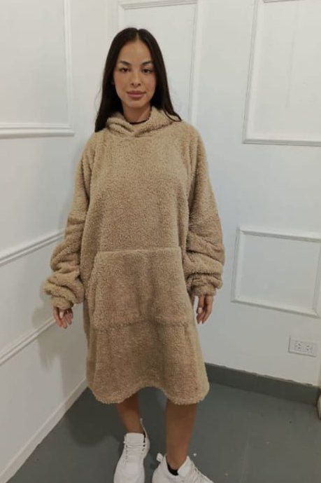 Mujer con un vestido extragrande de color bronce y capucha, de pie en una habitación y con zapatillas blancas.