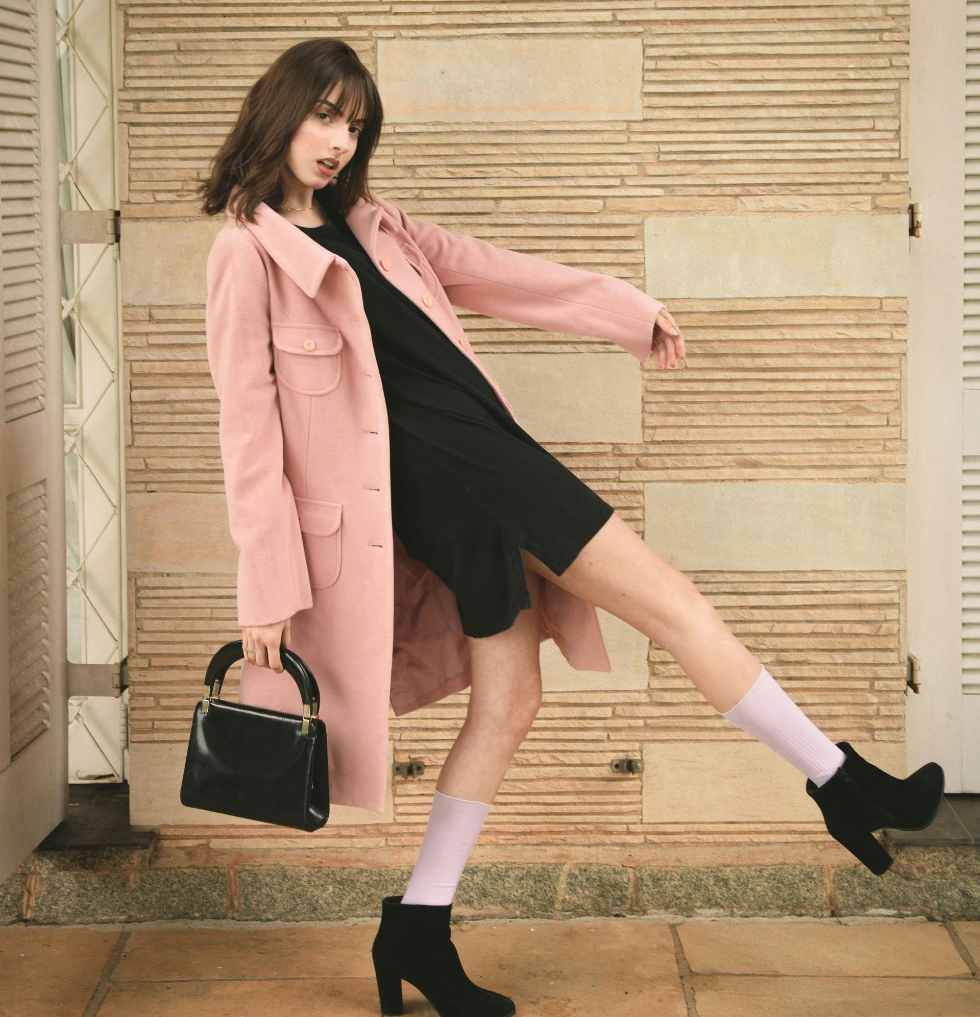 Mujer con abrigo rosa y vestido negro se balancea, sosteniendo un bolso, con botas negras y calcetines rosas.