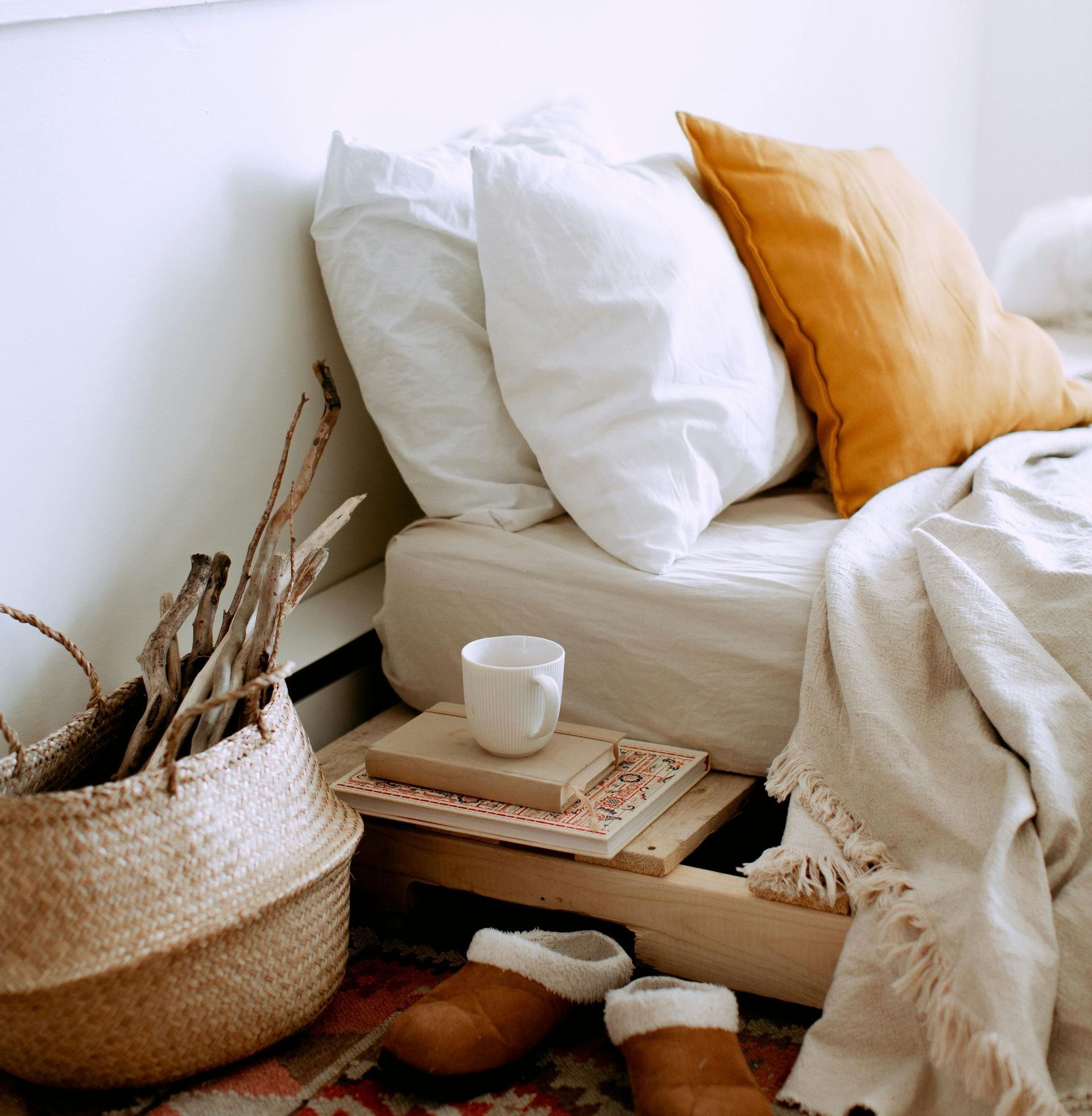 Rincón acogedor del dormitorio: cama con almohadas blancas y naranjas, cesta tejida con ramas y zapatillas marrones.