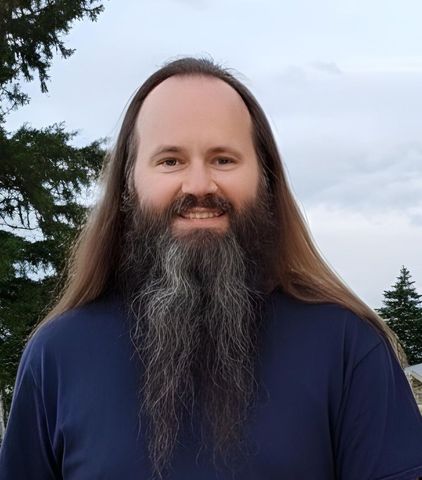 Shawn Aller with long brown hair and large graying beard smiles outdoors.