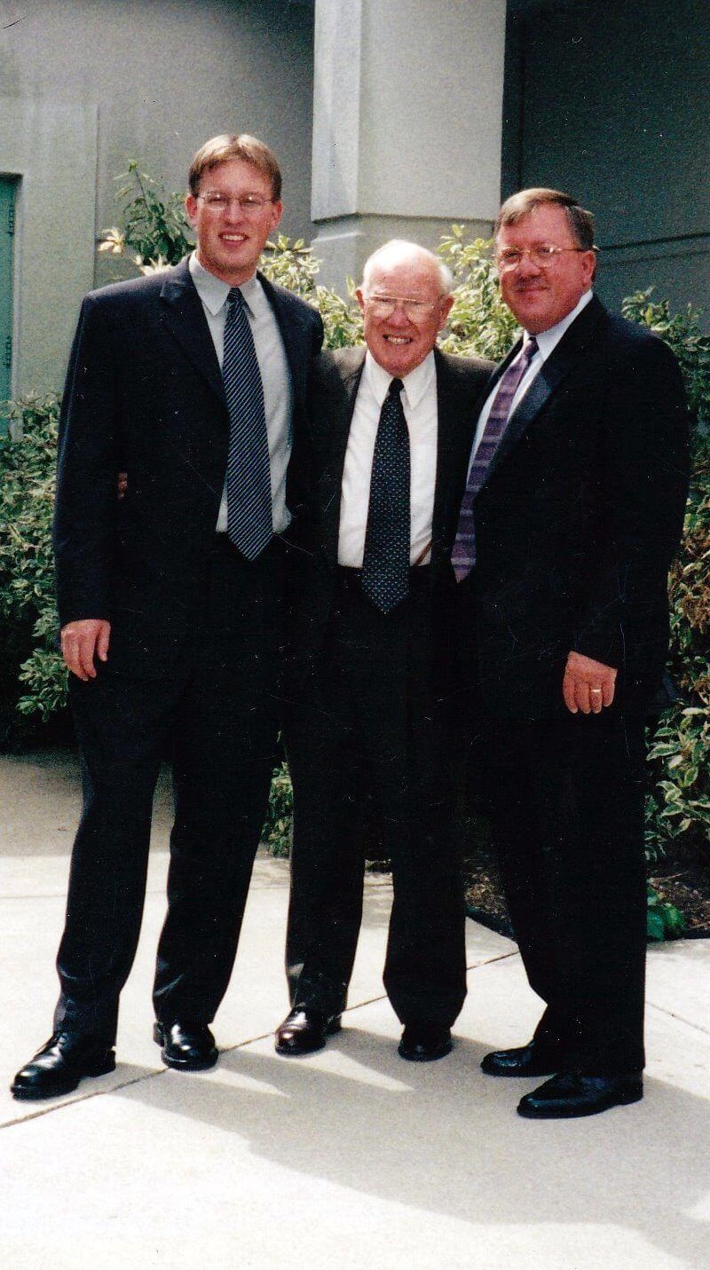 Three men in suits pose outdoors: a younger man, an older man, and a middle-aged man.