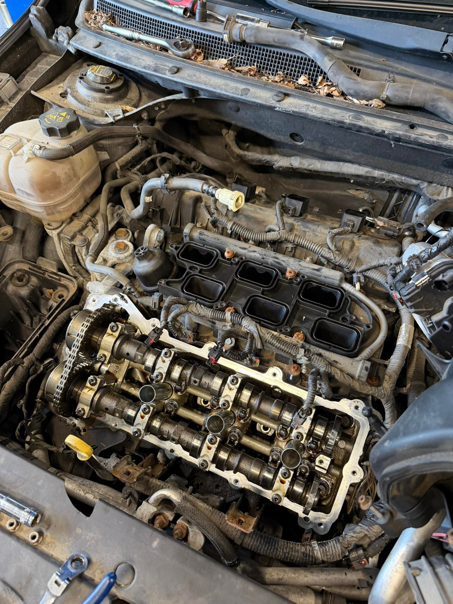 Car engine bay with the valve cover removed, exposing engine components and wiring during repair.
