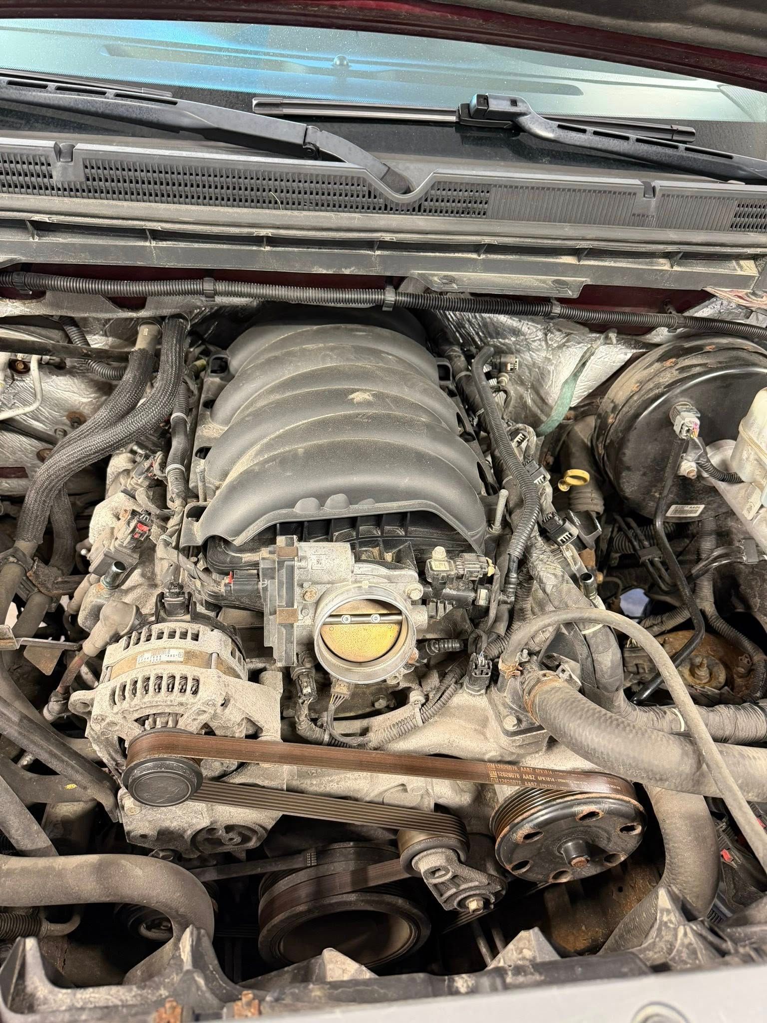 Car engine bay with exposed V8 intake manifold, belts, and hoses under the hood.