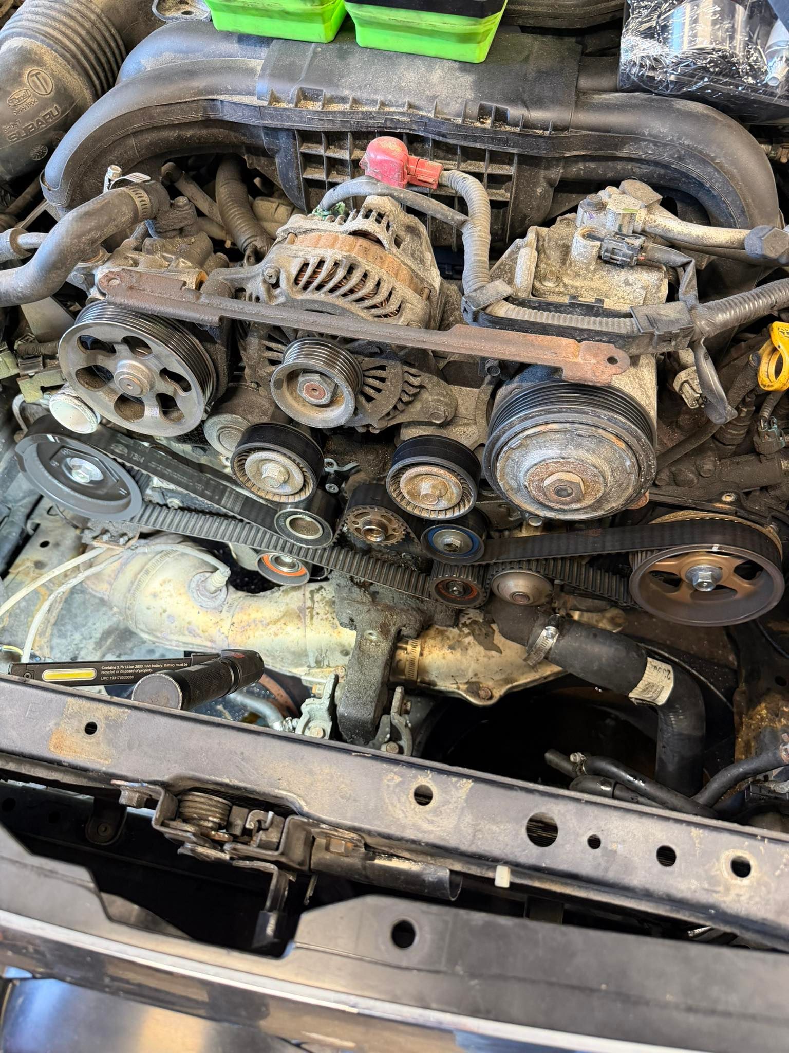Exposed car engine with serpentine belt, pulleys, and hoses in a dirty engine bay.