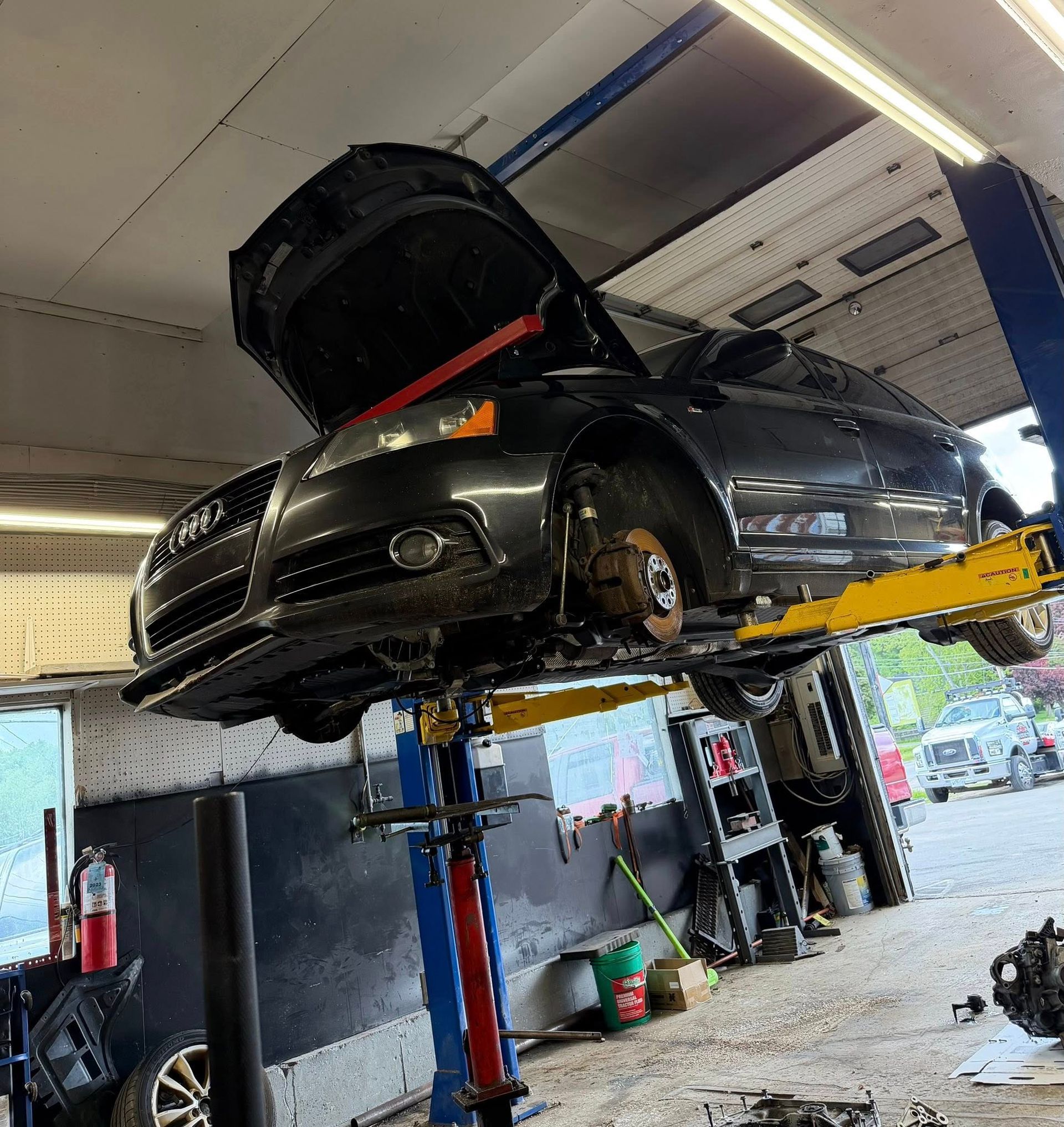 Black car lifted in an auto shop, hood open, with front wheel removed for repair.