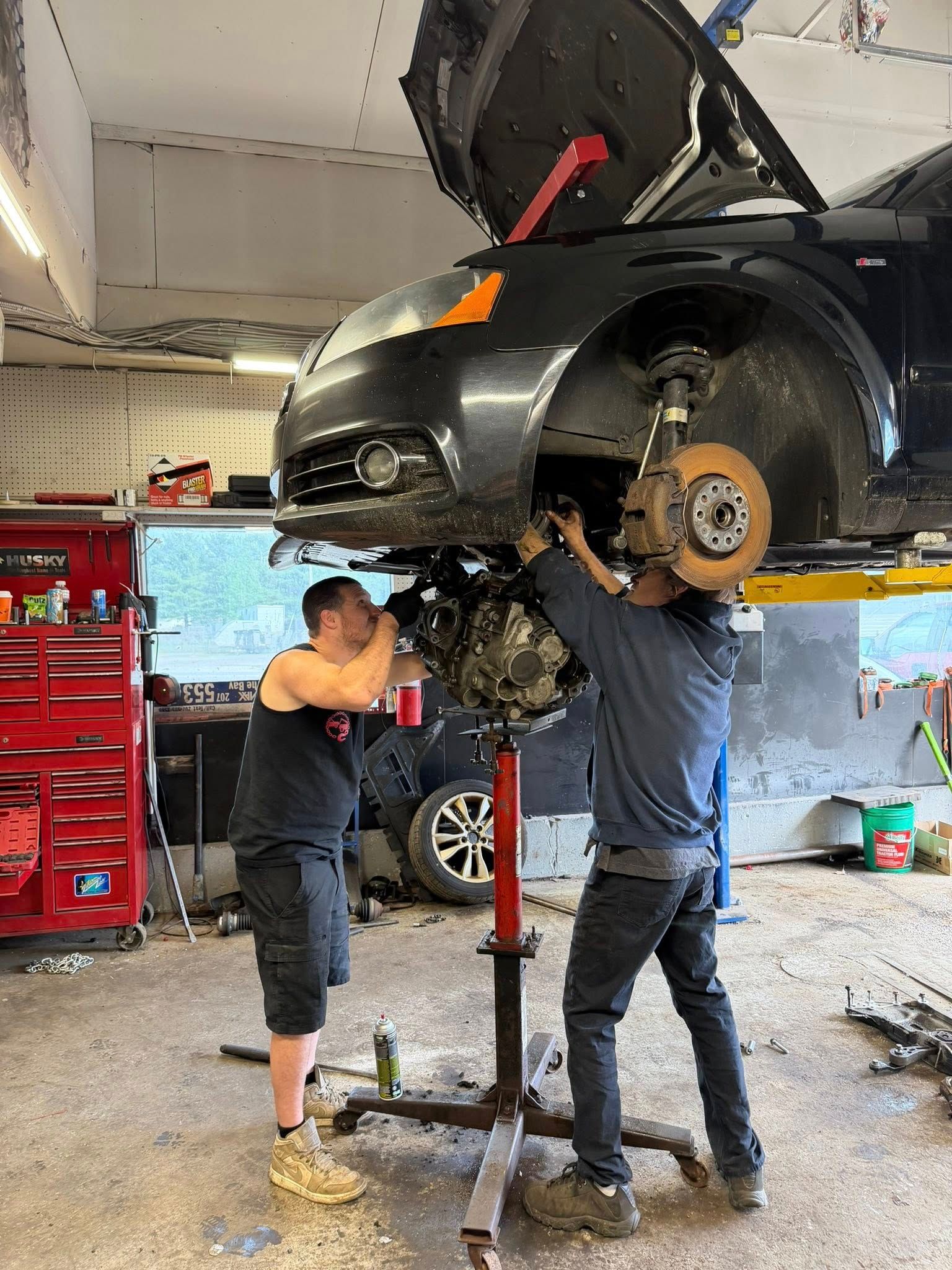 Two mechanics working under a raised black car in an auto repair garage
