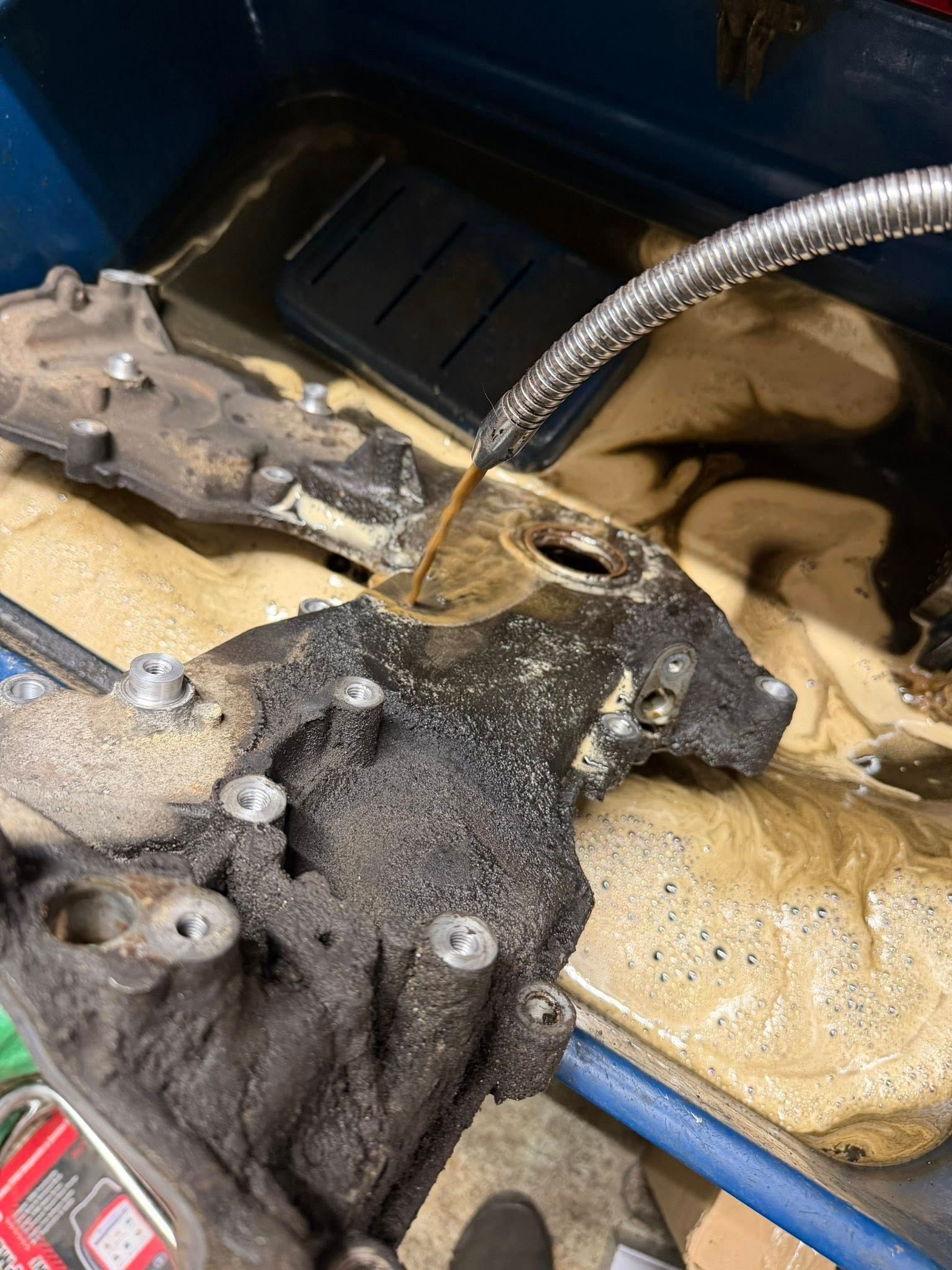 Mechanic’s engine bay with a greasy component, hose, and pooling brown liquid in a blue container