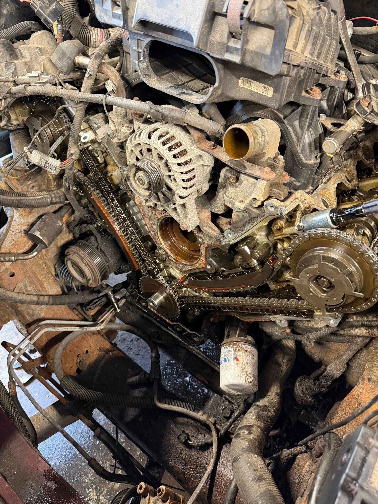 Severely damaged engine bay with exposed gears, hoses, and rusted mechanical parts in a scrapyard.