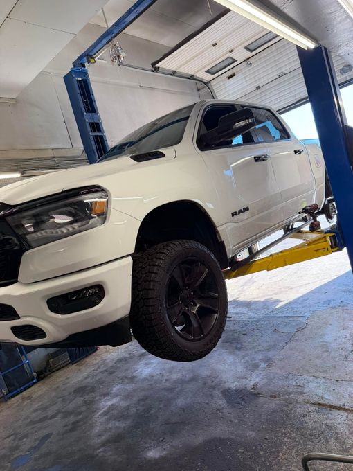 White pickup truck lifted on a car lift in an auto shop, with black wheels and open garage interior.