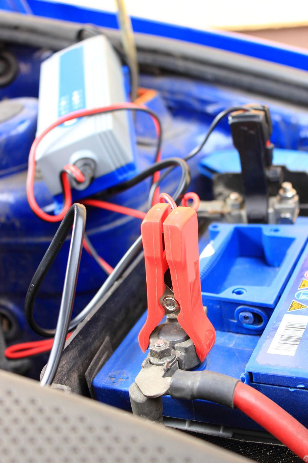 Red jumper clamp attached to a battery terminal with wires and a blue charger in the background