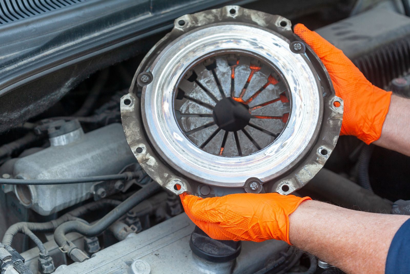 Gloved hands holding a car clutch assembly inside an engine bay