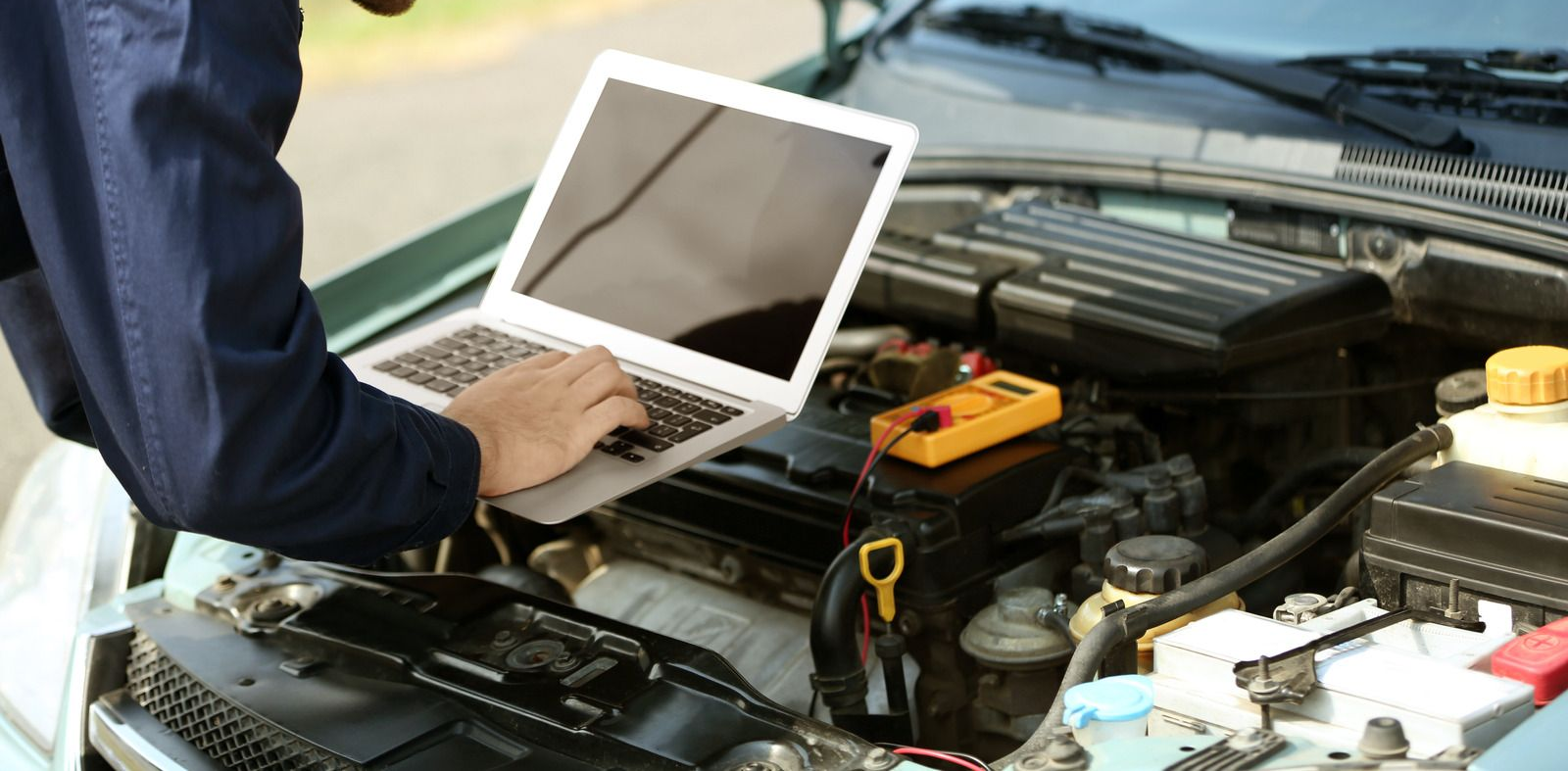 Mechanic using a laptop under an open car hood in a garage, with tools and engine parts visible.