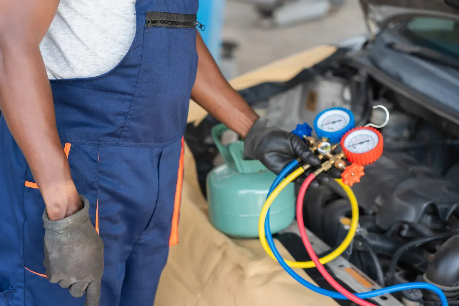 Mechanic holding AC manifold gauges beside an open car engine, with blue, yellow, and red hoses attached