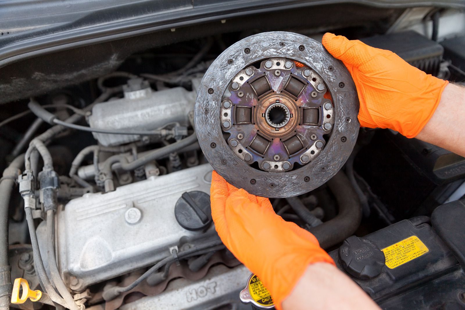 Hands in orange gloves holding a car clutch disc over an engine bay