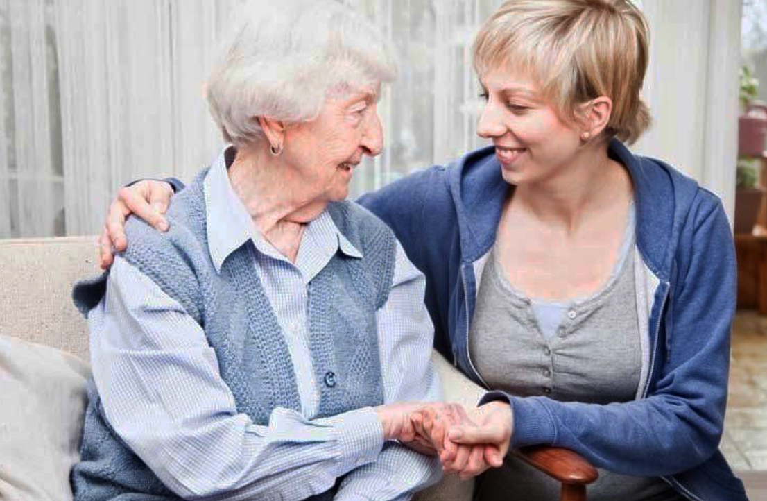 Eine Frau in einem blauen Pullover tröstet eine ältere Frau, die Händchen haltend auf einem Sofa sitzt.