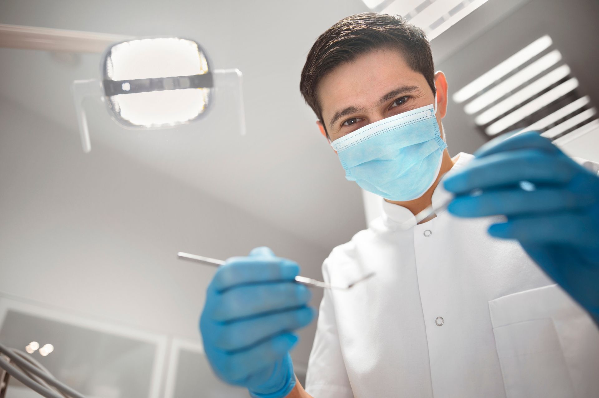 A dentist holds dental tools while wearing a mask, inside a dental office
