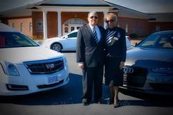 Man and woman pose between two cars outside a building with white columns. The man wears a suit, the woman a uniform.