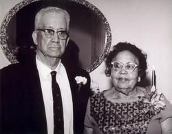 Elderly couple posing formally, man in suit with glasses, woman in patterned dress, mirror in background.