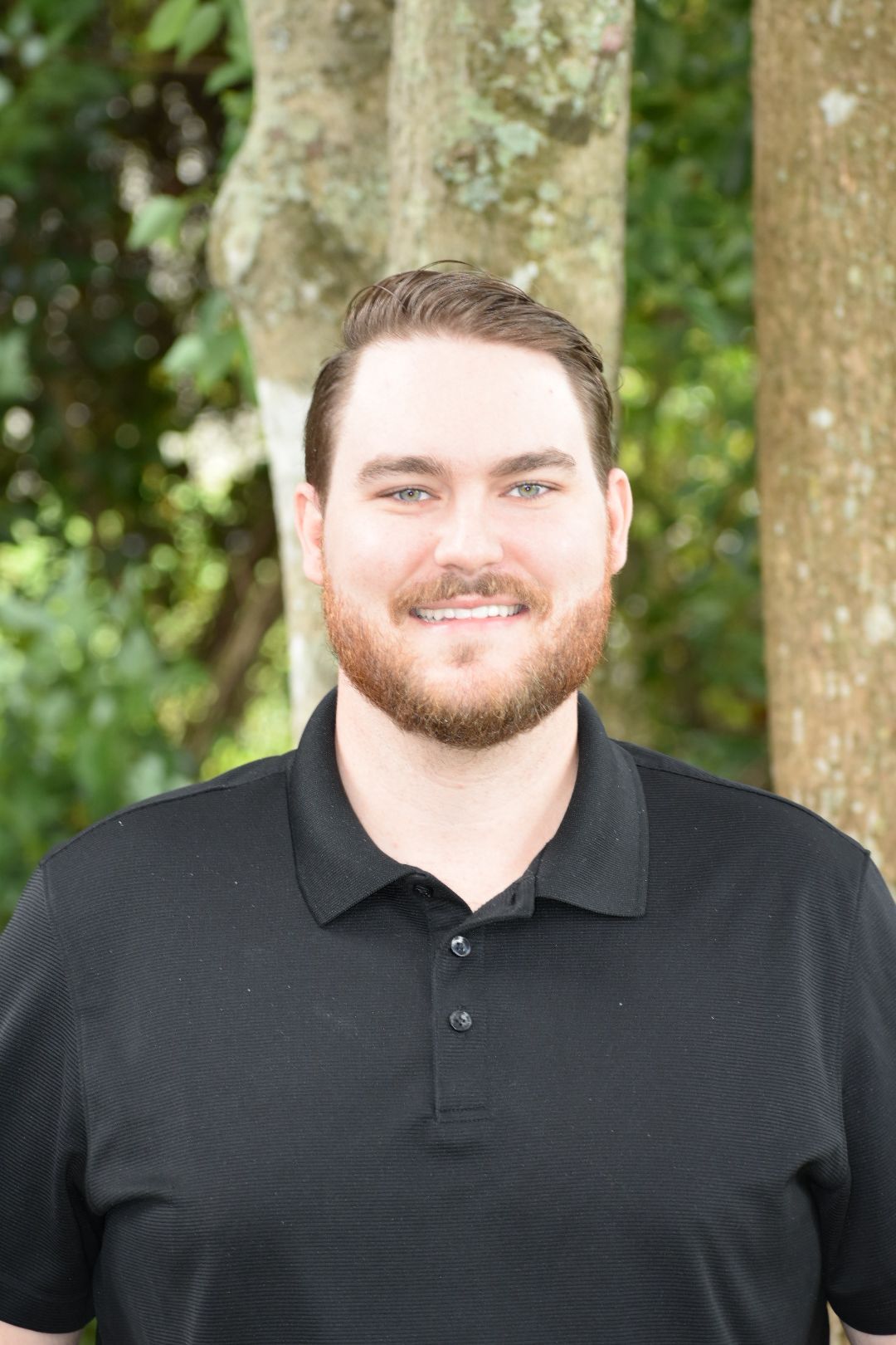 A man with a beard is wearing a black polo shirt and smiling for the camera.