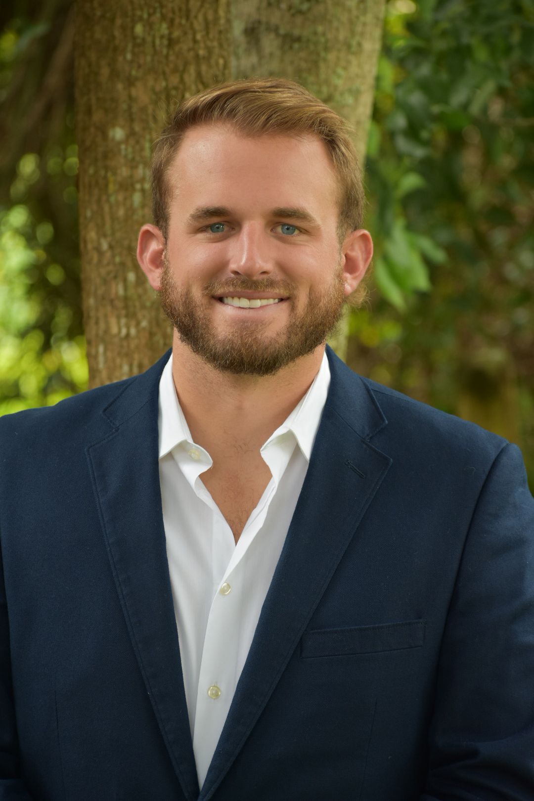 A man in a suit and white shirt is standing in front of a tree.