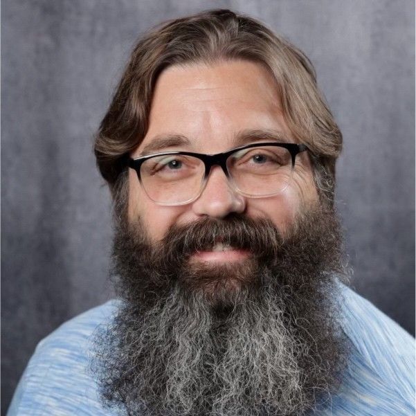 Man with glasses and a long beard smiling at the camera, blue shirt, grey background.