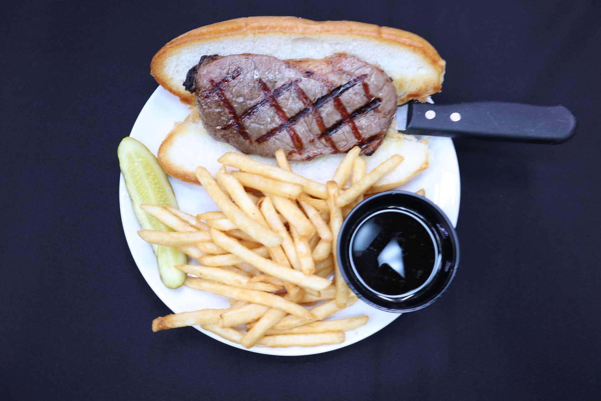 Steak sandwich with fries, pickle, and dip on a white plate.
