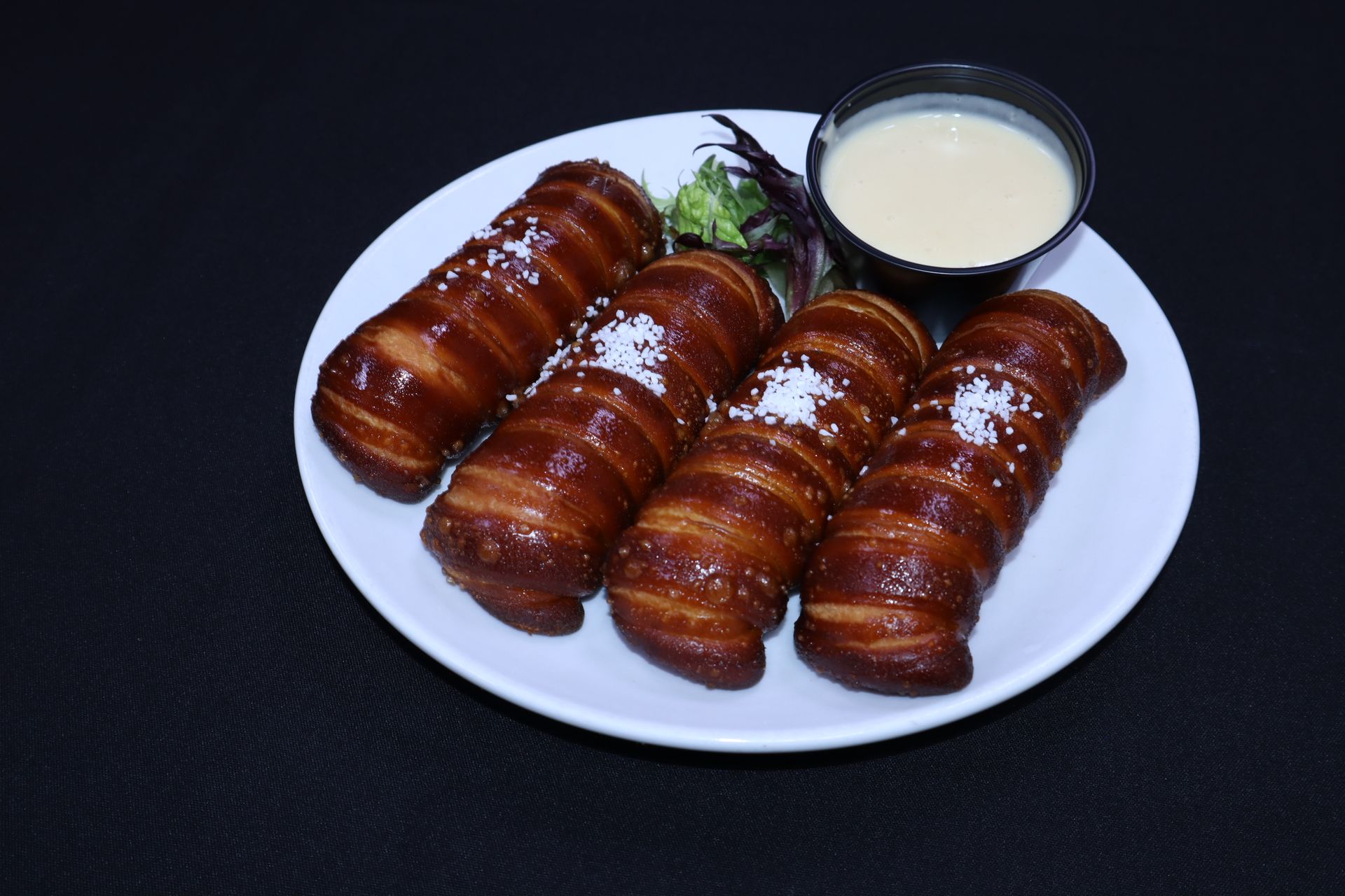 Four pretzel-wrapped sausages on a white plate with a dip and greens on a black background.