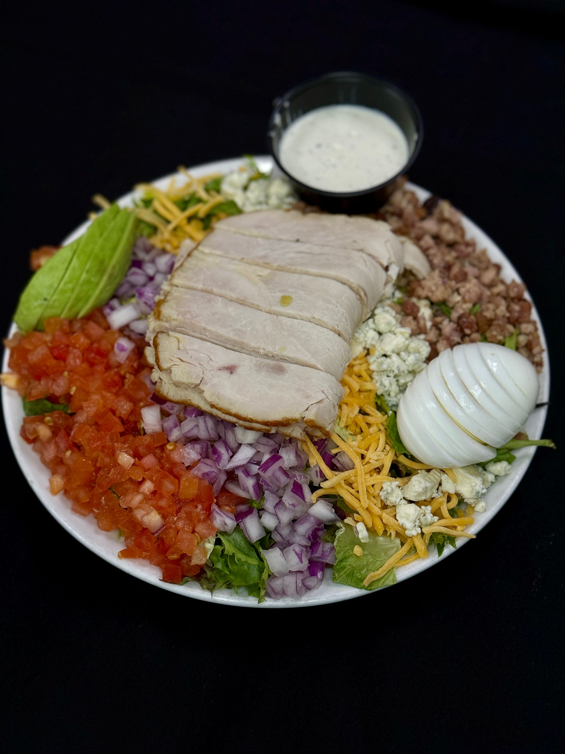 Salad with turkey, avocado, tomato, egg, and blue cheese, with a side of dressing on a white plate.