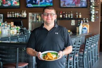 Man in black shirt holding plate of food, standing in a restaurant with a bar in the background.