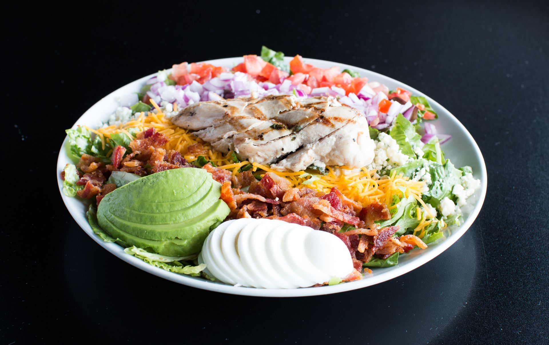 A salad with avocado, bacon, cheese, tomatoes, and onions on a plate.