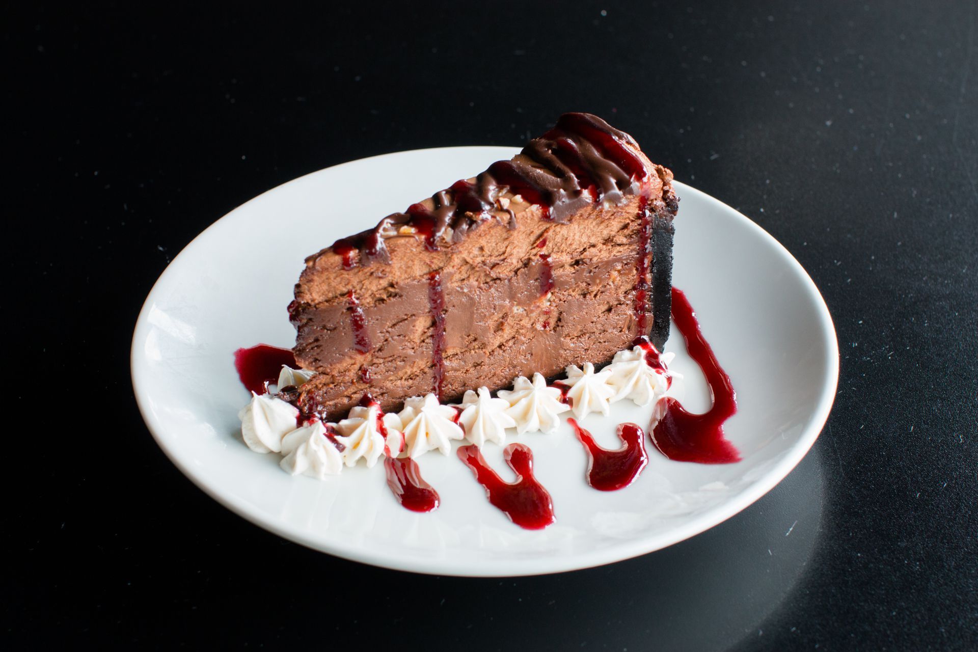 A slice of chocolate cheesecake with whipped cream and raspberry sauce on a white plate.
