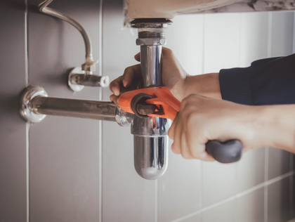 Hands using an orange wrench to tighten a chrome pipe under a bathroom sink against a tiled wall.