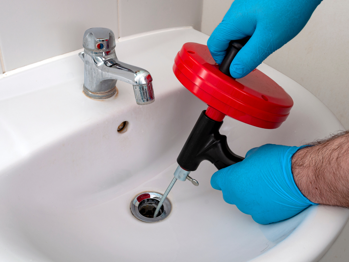 Hands with blue gloves using a drain snake in a white sink to unclog a drain.