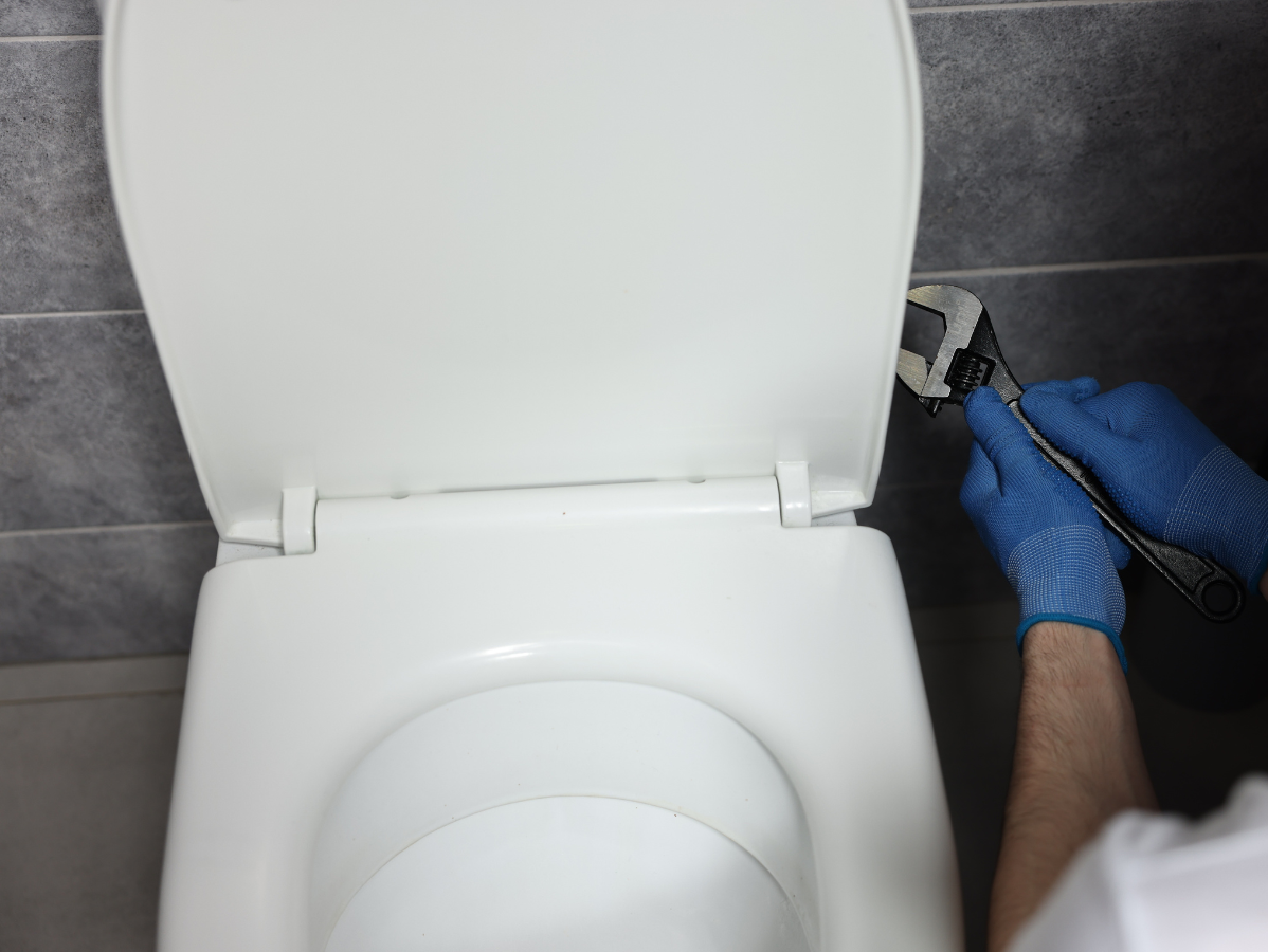 Person using a wrench to repair a toilet seat. They are wearing blue gloves in a bathroom.