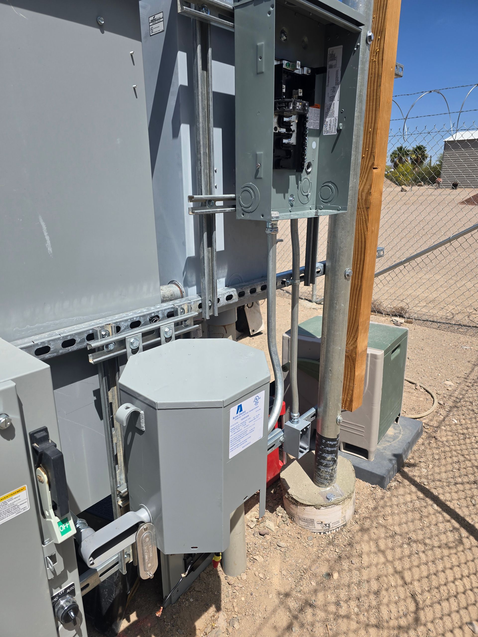 Electrical equipment mounted outdoors with conduit, on a concrete base near a fence.