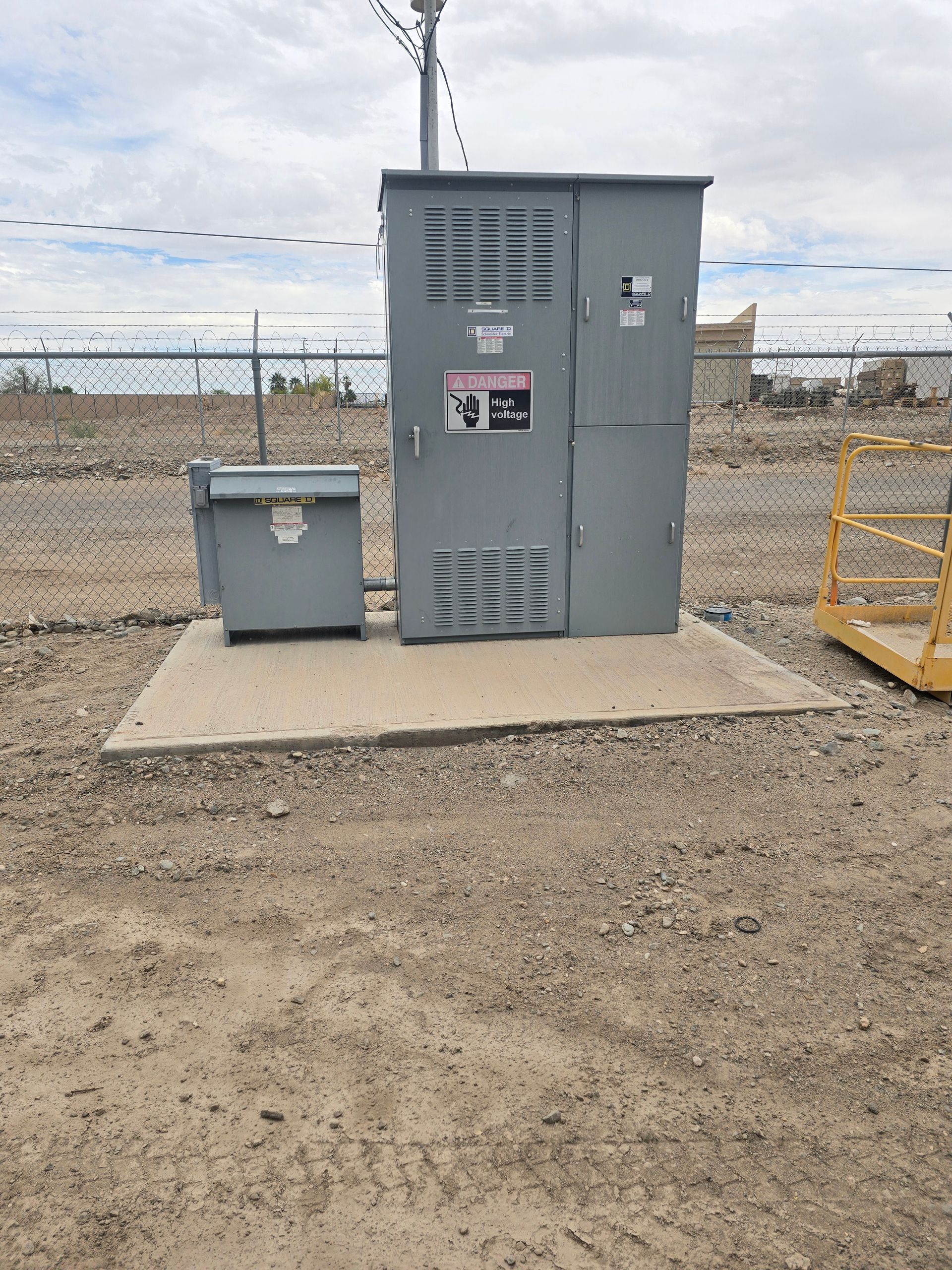 Gray electrical boxes on a concrete pad, outdoors, near a fence, and telephone pole.