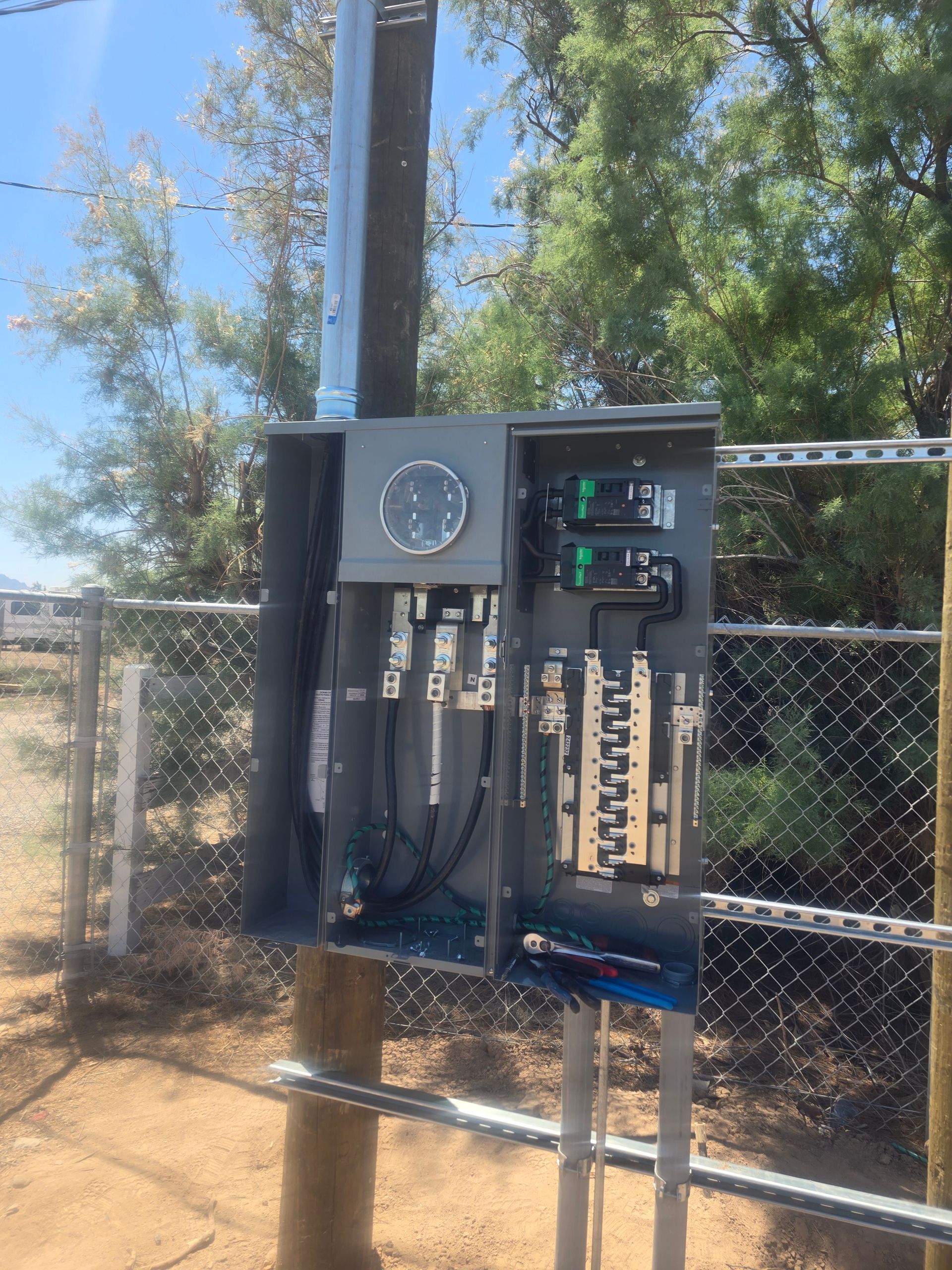 Electrical box mounted on a pole, containing meters and circuit breakers, near a fence and trees.