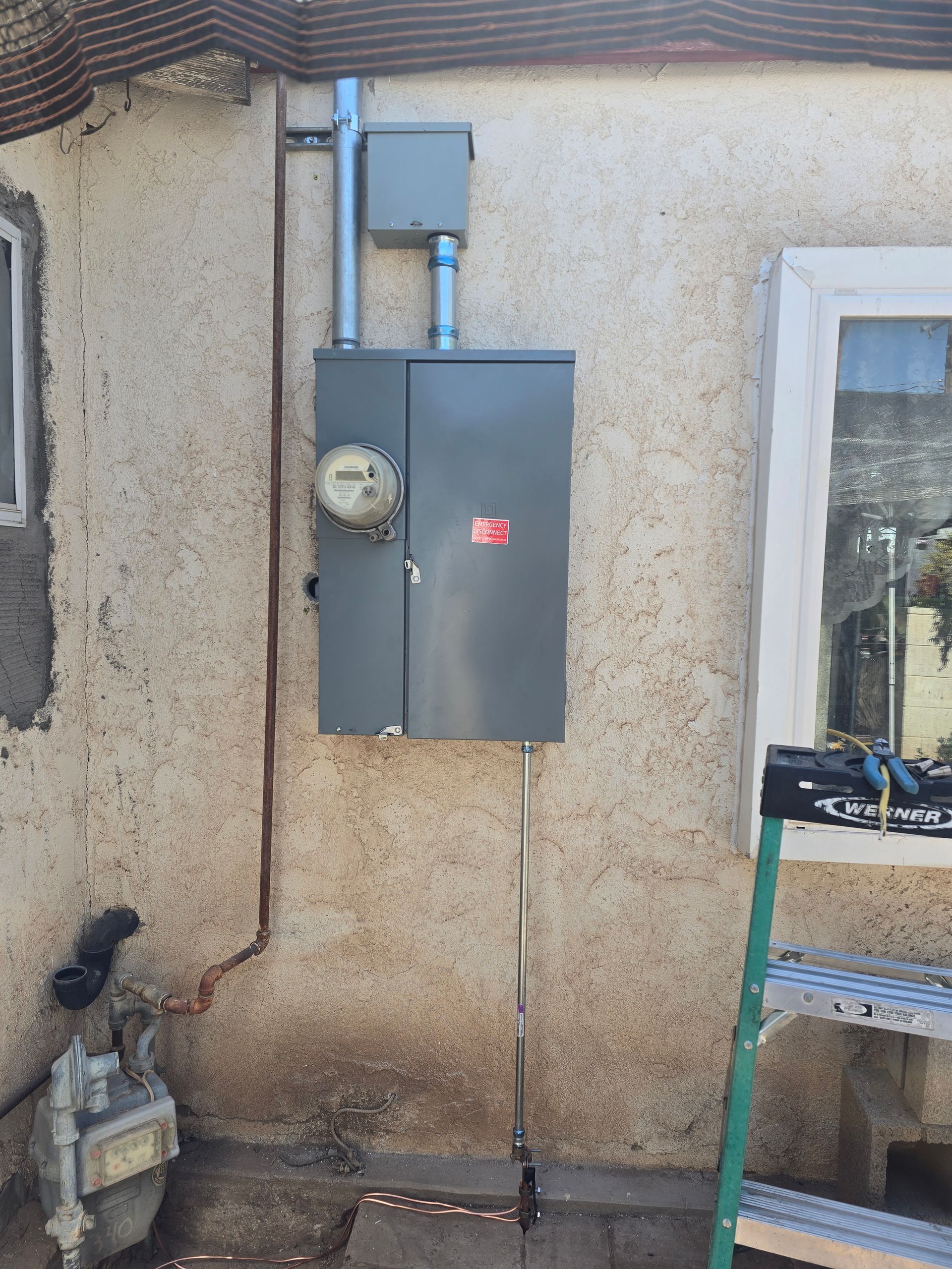 Electrical panel and meter on a stucco wall next to a window and gas meter. A ladder is nearby.