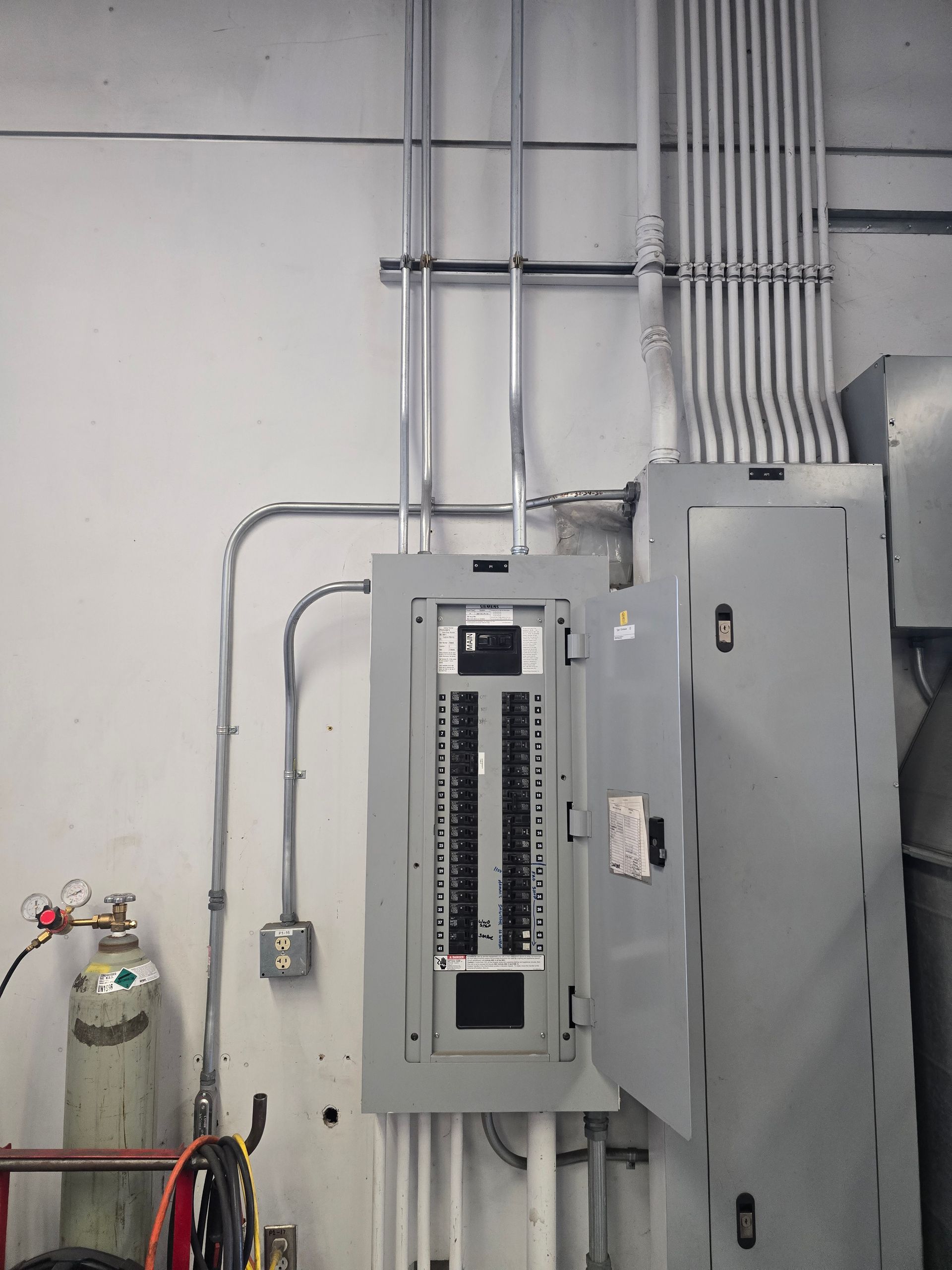 Electrical panel with conduit and wiring, mounted on a white wall in a commercial setting.