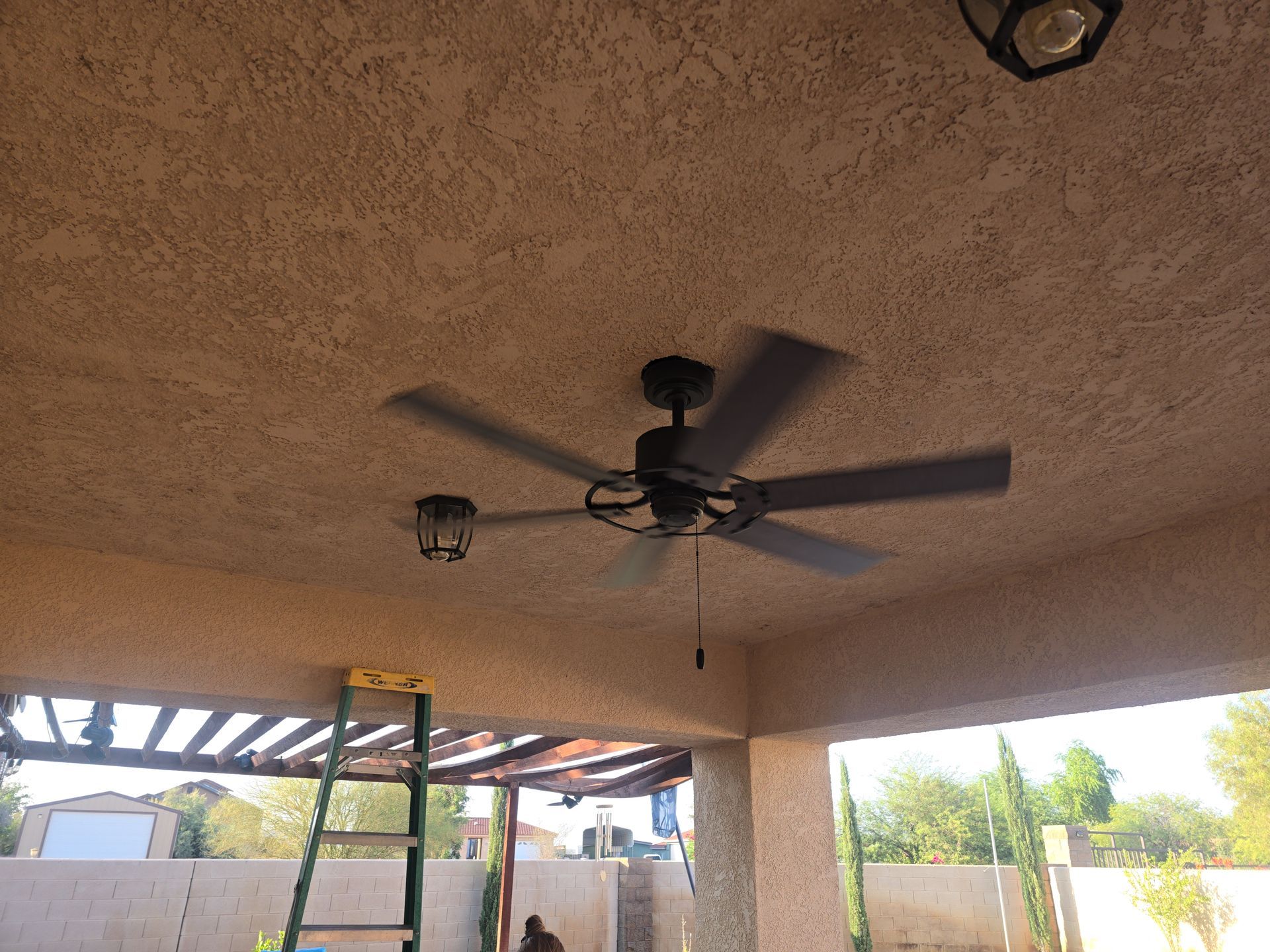 Ceiling fan and lights on a stucco patio ceiling with a ladder.