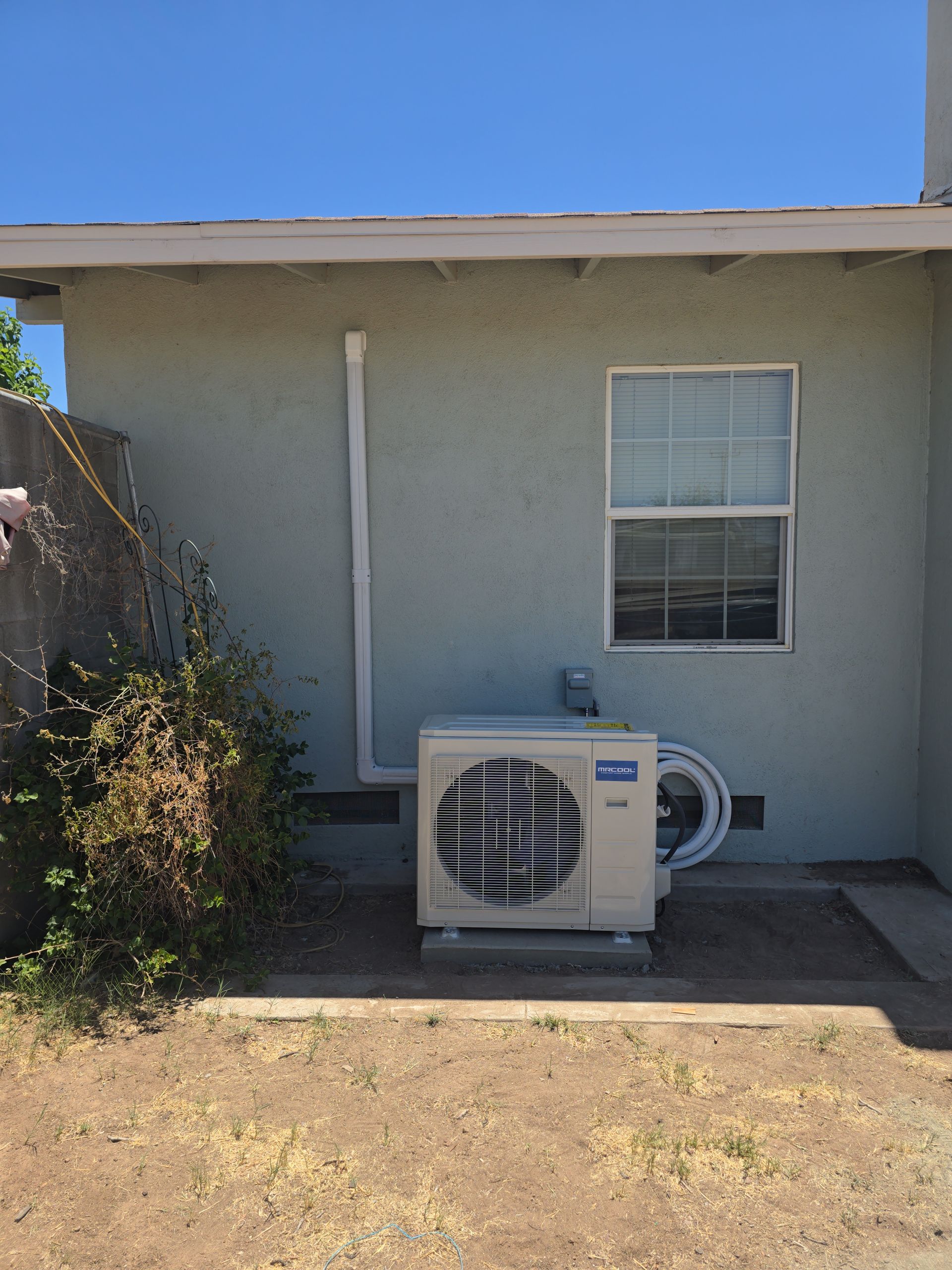 An outdoor air conditioning unit sits on a concrete pad near a building's pale green wall, below a window and white pipe.