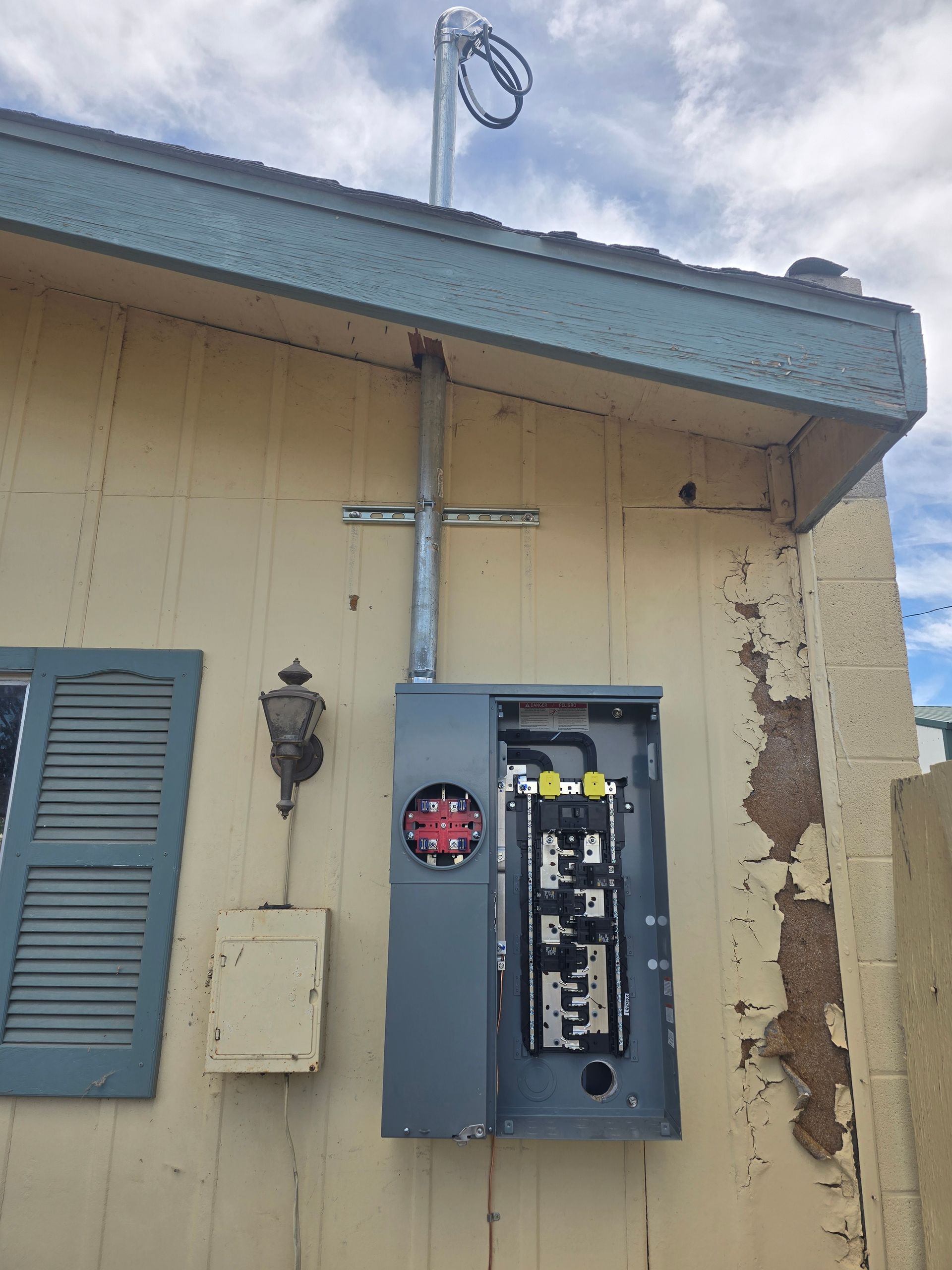 Gray electrical panel mounted on a yellow building exterior with conduit running to a pole-mounted utility wire.