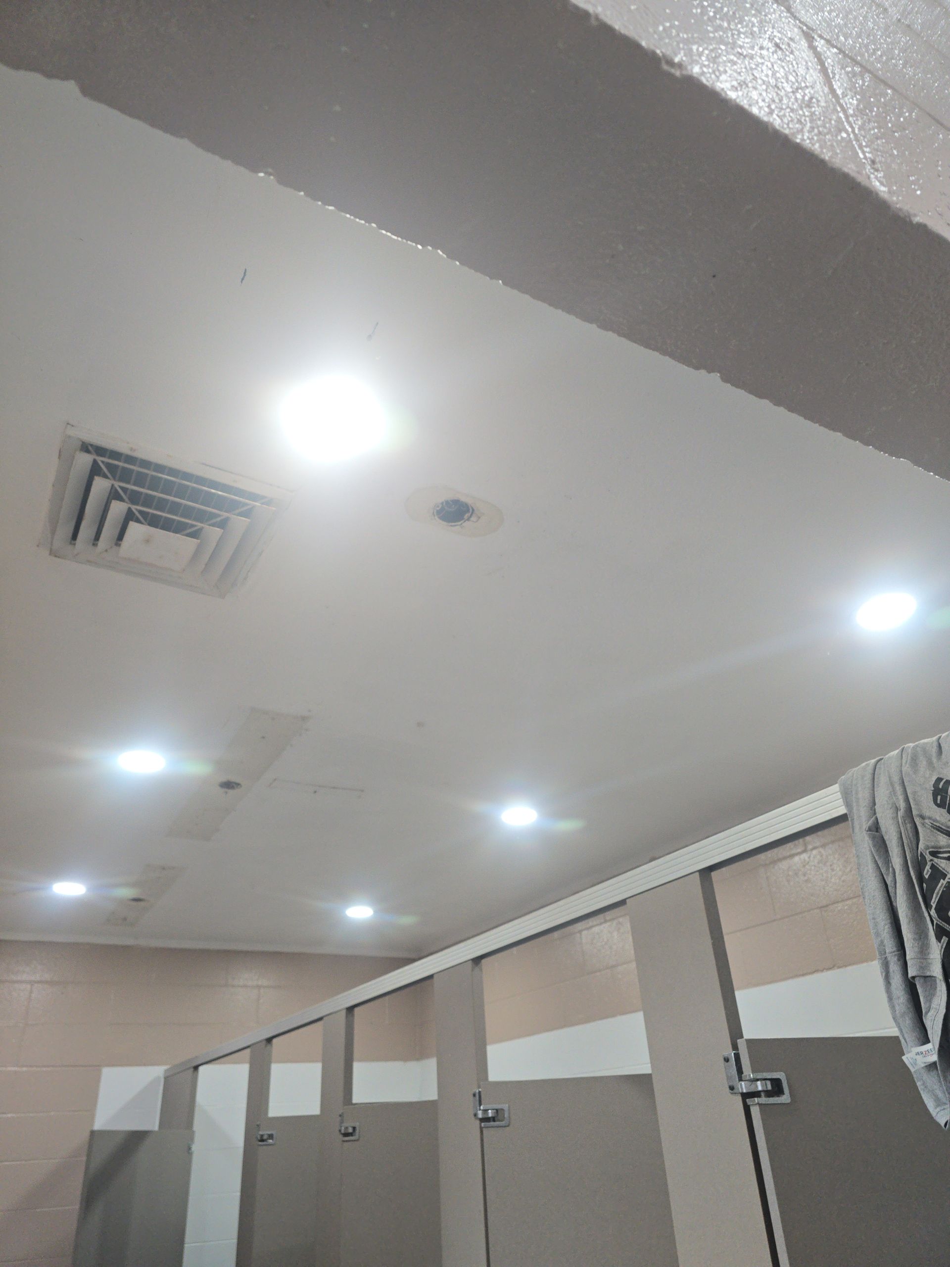 Public restroom ceiling with recessed lights, vent, and stall dividers.