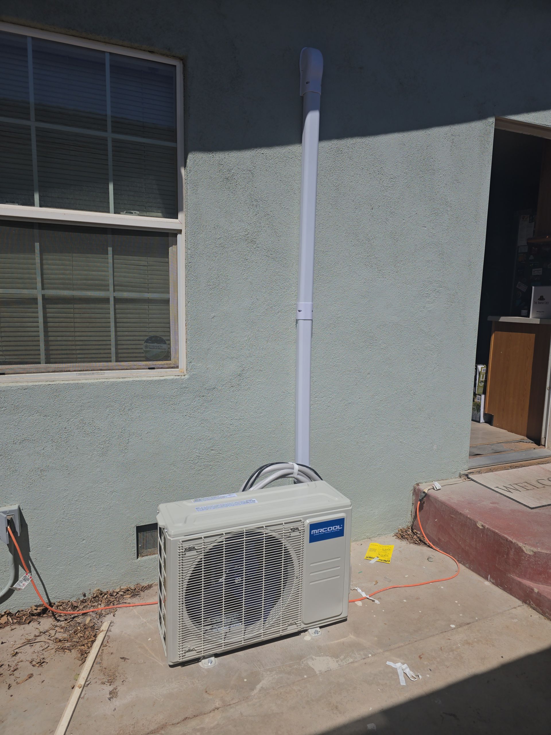 Air conditioning unit attached to a building, with a vent pipe running upwards. Gray stucco wall, window, and concrete.