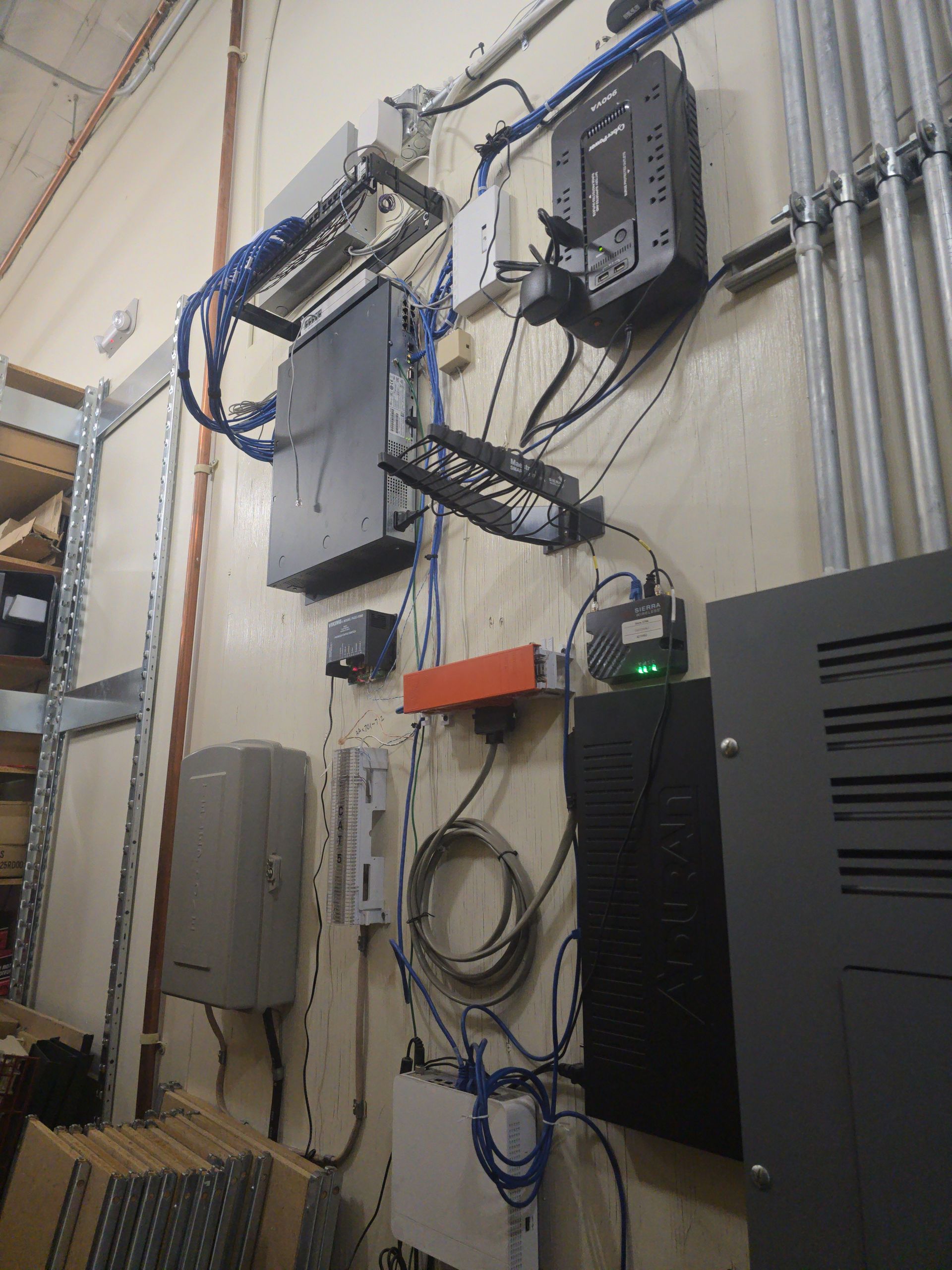 Network equipment mounted on a wall with cables. Various black, gray, and orange boxes and panels.